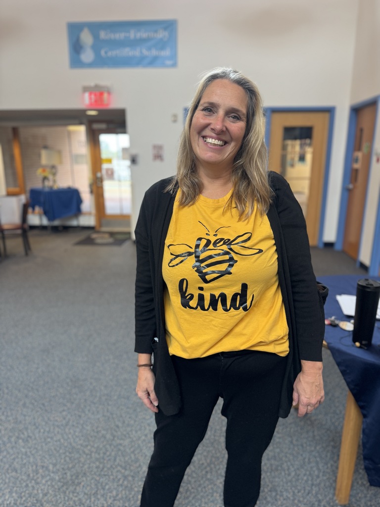 A woman with blond hair smiles in a yellow shirt that says "Be Kind" and black pants.