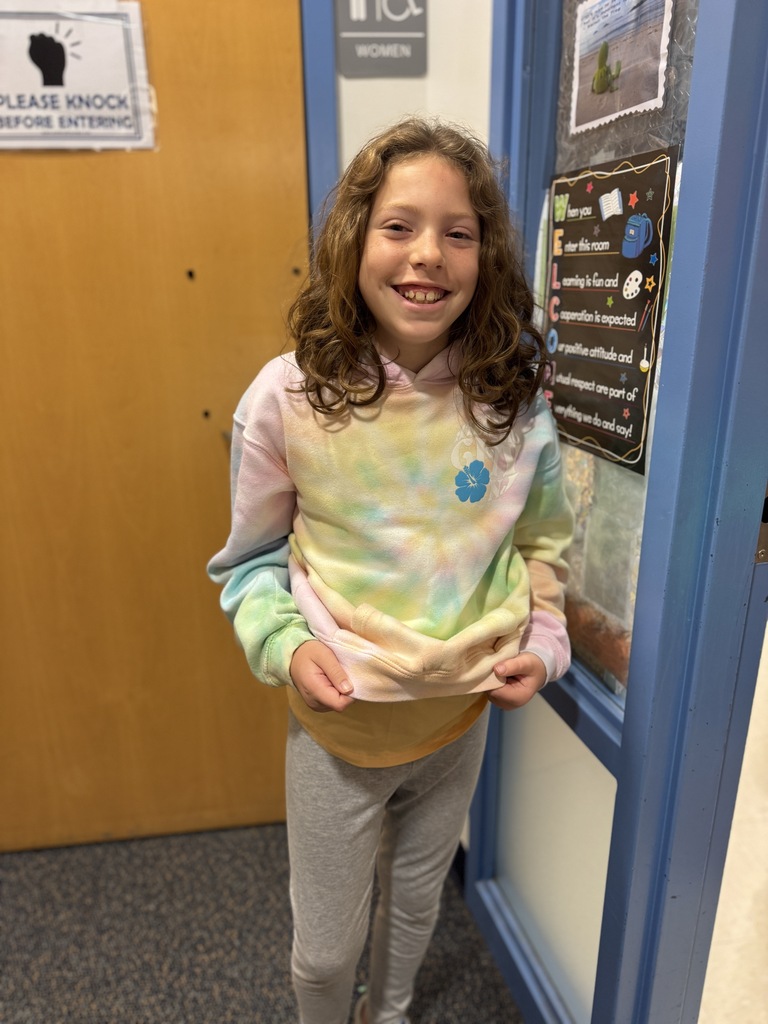 A girl in a tie-dye sweatshirt with a flower smiles and stands in a hallway.