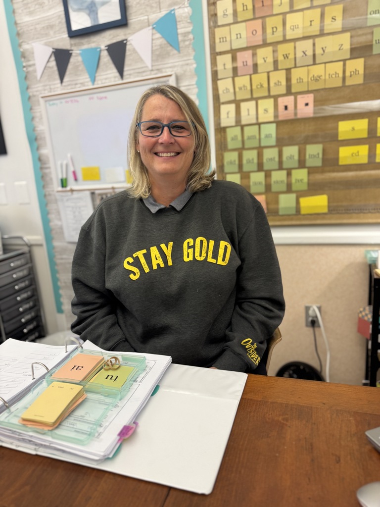 Woman in glasses and a black sweatshirt reading "Stay Gold" sits at a desk with a binder.