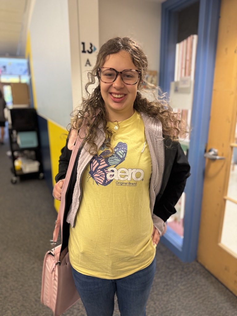 Woman with curly hair and glasses stands in hallway with yellow shirt, black jacket, jeans, and pink purse. Behind her, doors and yellow and blue wall.