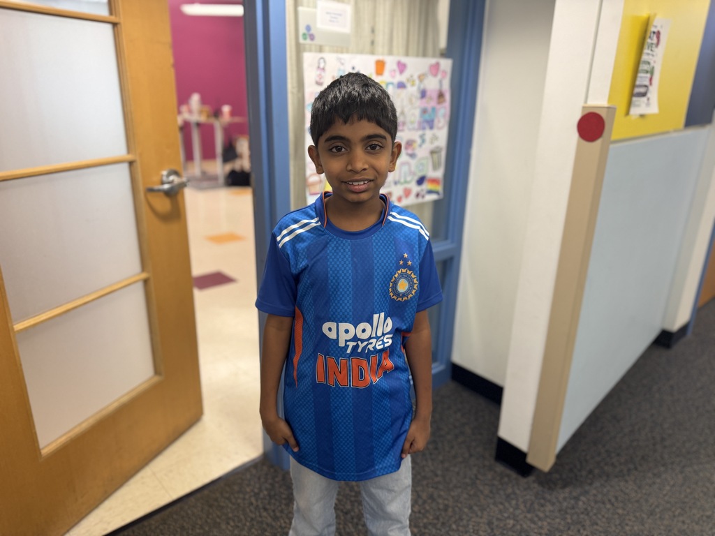 A young boy in a blue soccer jersey stands in a hallway with a door and bulletin board behind him.