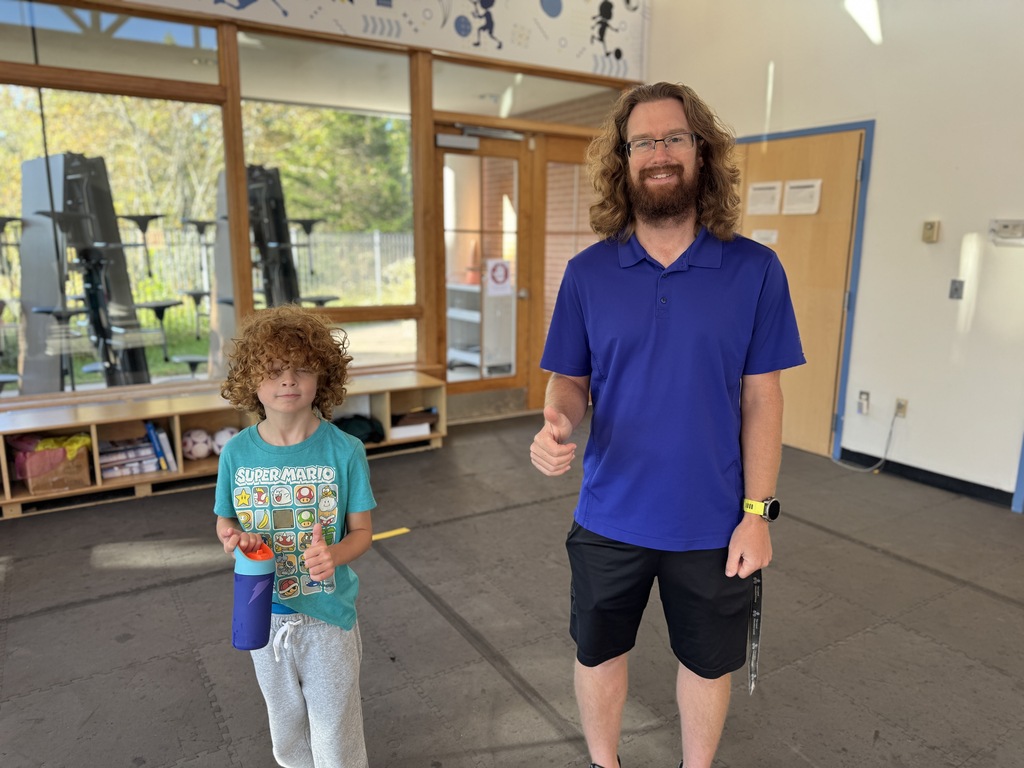 A man and child stand in a gym, smiling. The man wears a blue shirt and black shorts. The child holds a bottle. Behind them are benches and equipment.