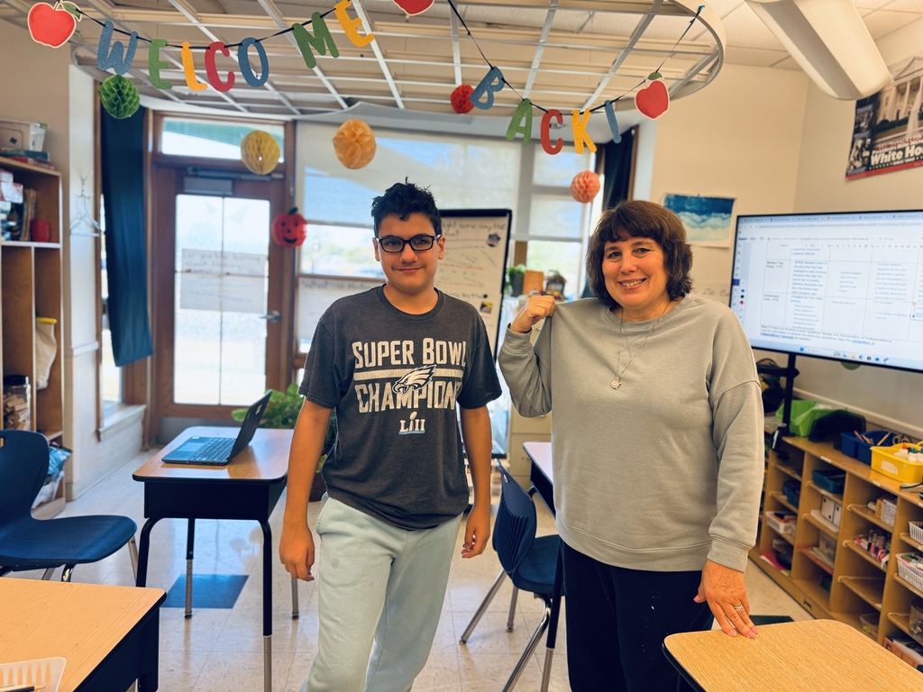 A man and a woman standing in a classroom. Behind them is a bulletin board and a large screen.