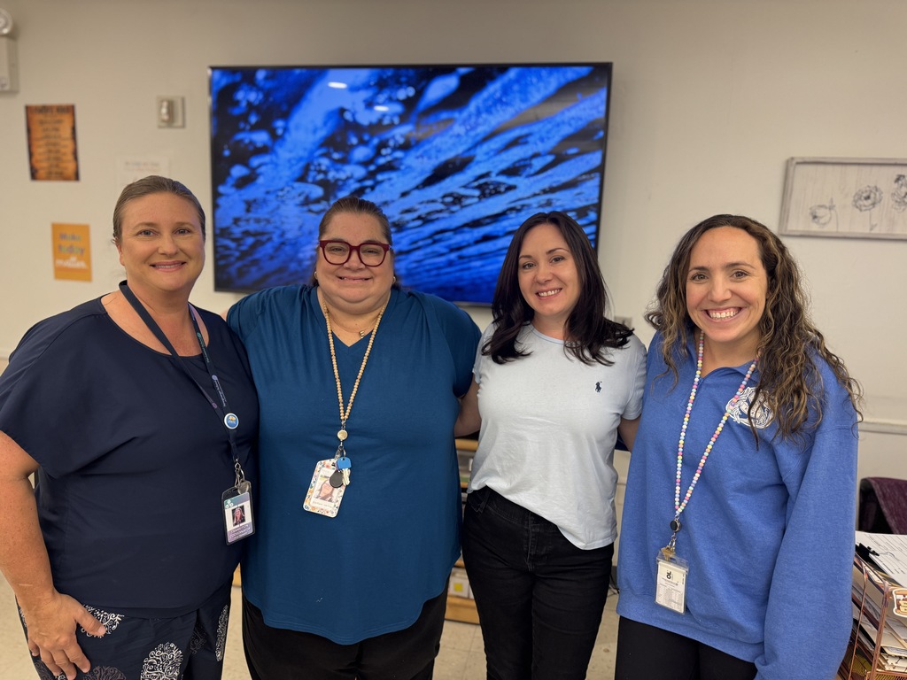 Four women stand together indoors. One wears a blue shirt, and three wear white shirts. A large blue screen is mounted on the wall behind them.