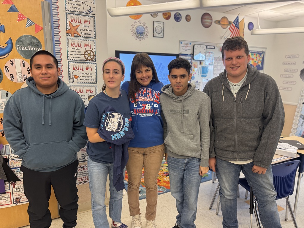 Four people stand together in a classroom. A man and woman wear blue hoodies, and two men wear gray hoodies.