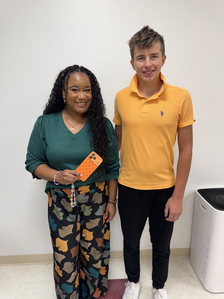 Two people stand against a white background, one holding an orange phone case and smiling. A person wears a yellow shirt, black pants, and white shoes.