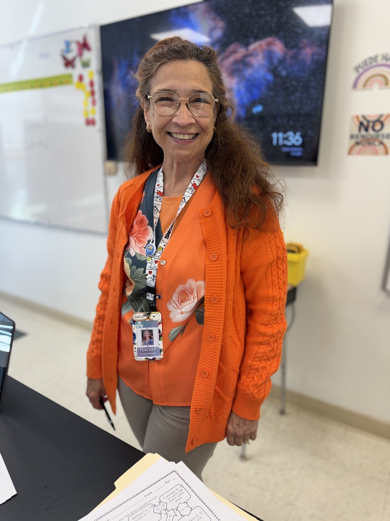 A woman wearing an orange cardigan and glasses stands in a classroom, smiling and facing the camera.