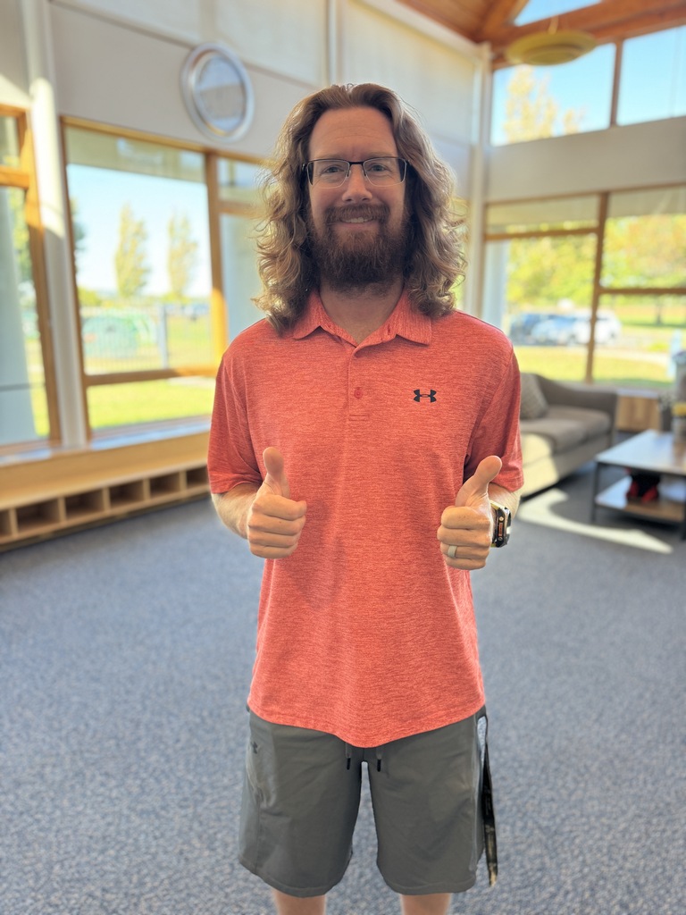 Man with long hair and beard, wearing orange polo shirt, giving thumbs up. Behind him, a couch, table, and large windows.