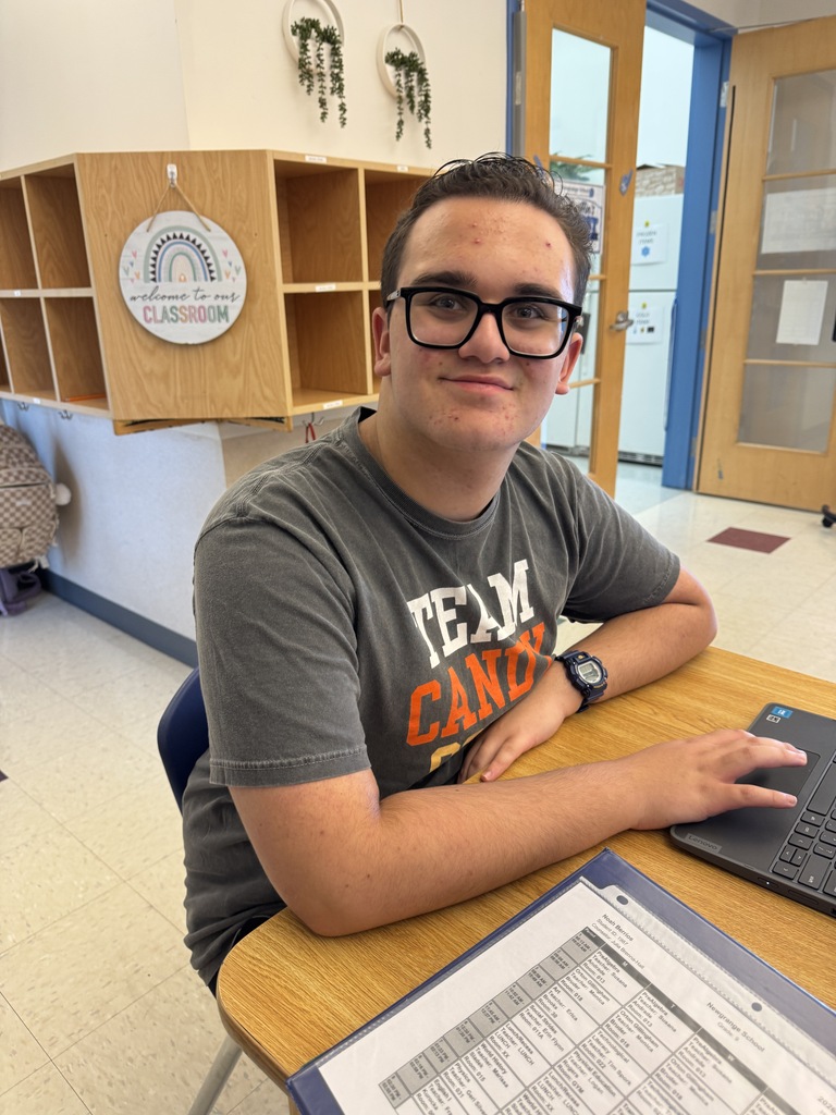 A man sits at a desk, wearing glasses and a gray T-shirt. He smiles while using a laptop.