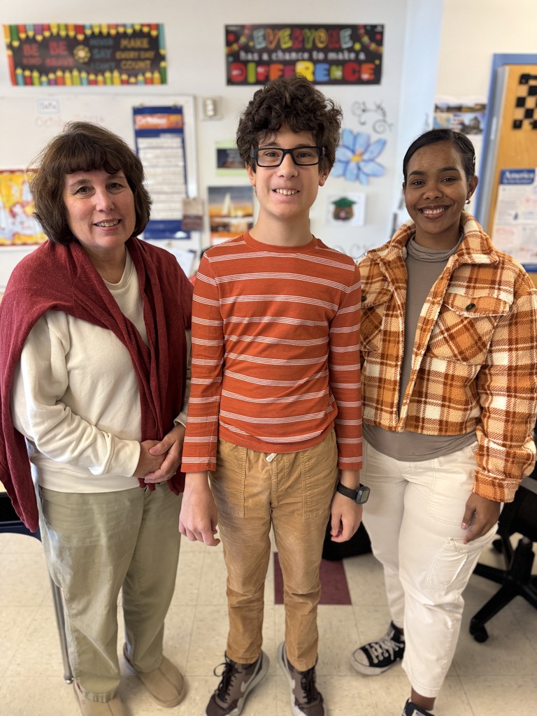 Three people stand in a classroom. A woman wears a red shawl. A boy has glasses and a striped shirt. Another girl has a plaid jacket.
