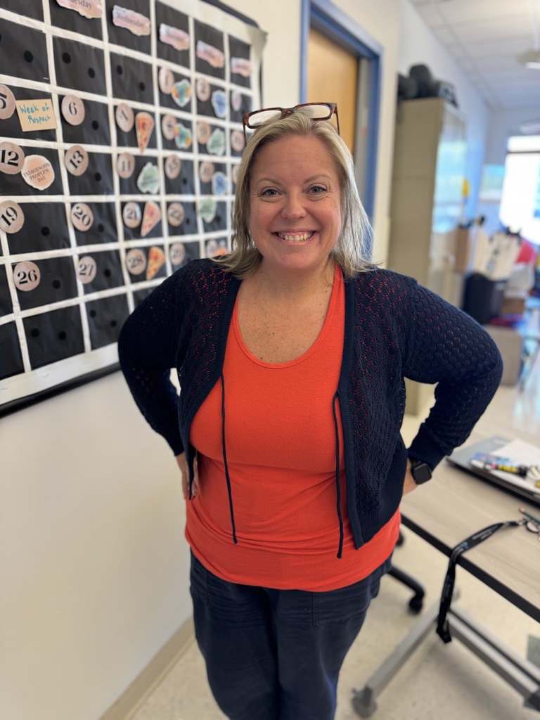 Woman with short hair wearing orange tank top and blue cardigan stands with hands on hips.