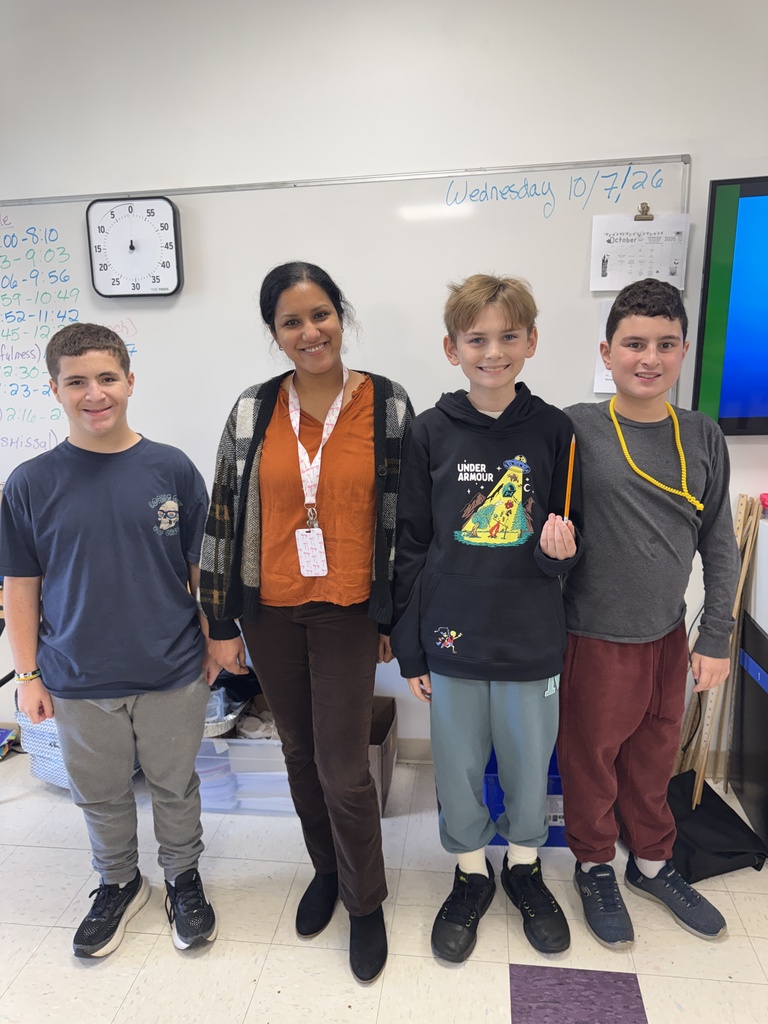 Four children and an adult stand in a room. Three boys wear hoodies and sneakers, and the woman wears a scarf. A clock and a whiteboard are in the background.