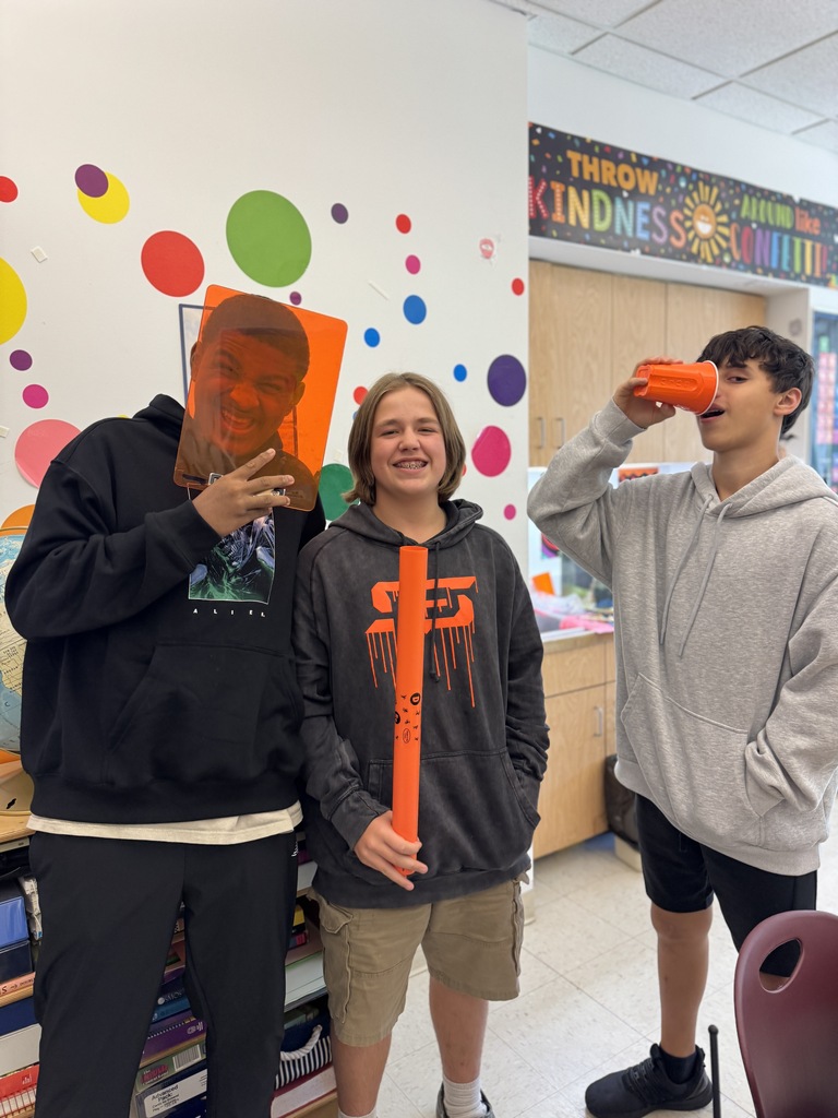 Three individuals in a classroom with a colorful wall. One holds a large photo, one holds a red cup, and the third holds a long orange object.