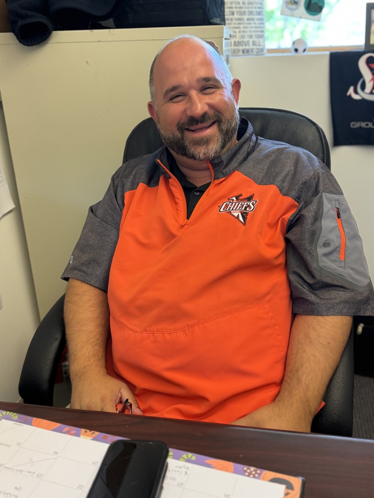 A man wearing an orange and gray jacket sits at a desk, smiling. An open binder and cell phone are on the desk. A wall with a logo is behind him.
