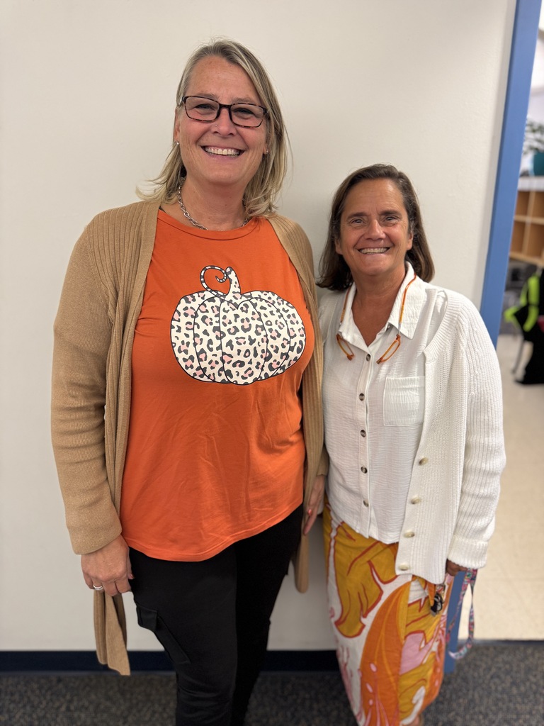 Two women smile and stand next to each other. One wears a pumpkin-printed orange shirt, and the other wears a white cardigan.