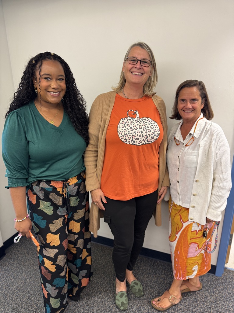 Three women in a room, one with glasses, one in an orange shirt, and one in a green shirt.