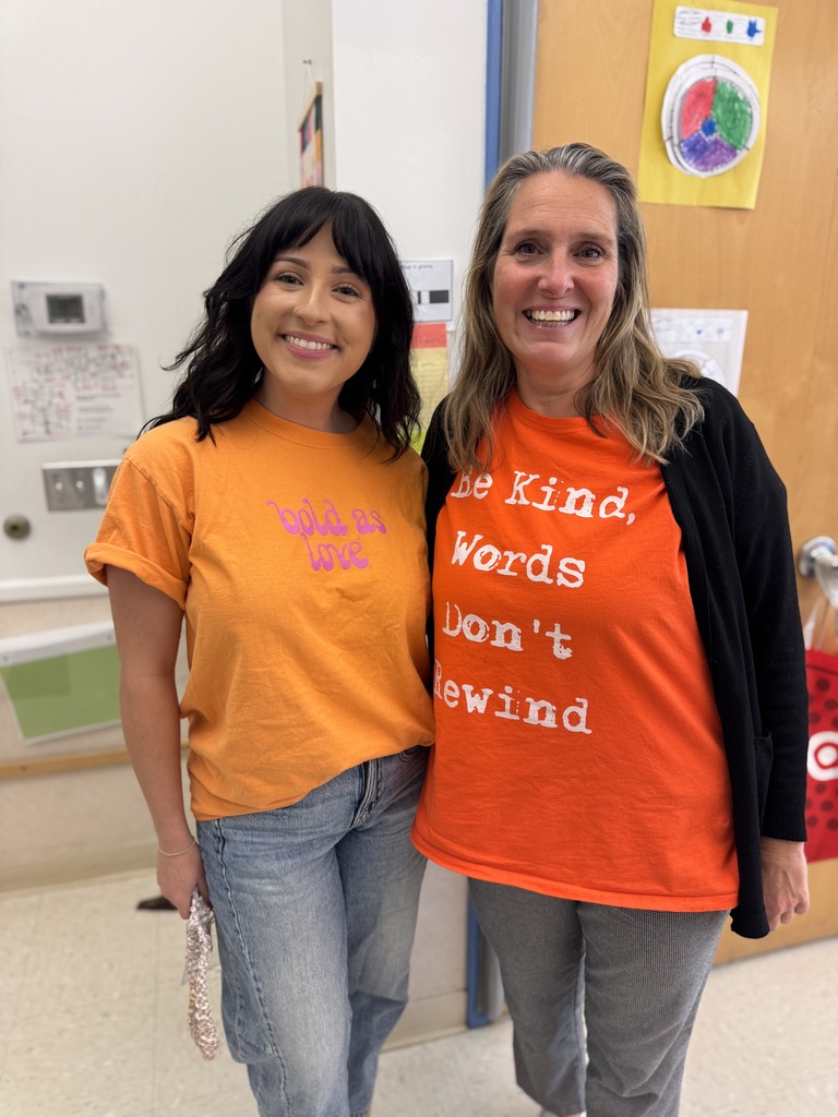 Two women in an indoor hallway, smiling. One wears an orange t-shirt, the other an orange shirt with black sleeves. Behind them is a brown door and a bulletin board.
