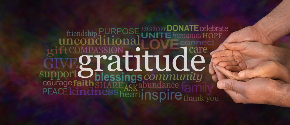 A word cloud anchored by the word 'gratitude' alongside several hands cupping and supporting each other against a dark, colorful background.