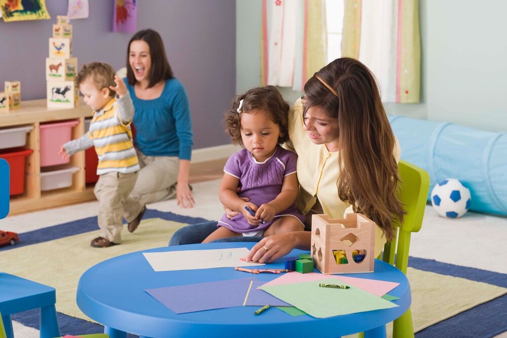 A teacher helping a young child color with crayons in a daycare classroom.