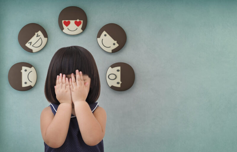 A child with short dark hair covers their face with their hands, standing in front of a wall displaying five cartoon faces showing different emotions.
