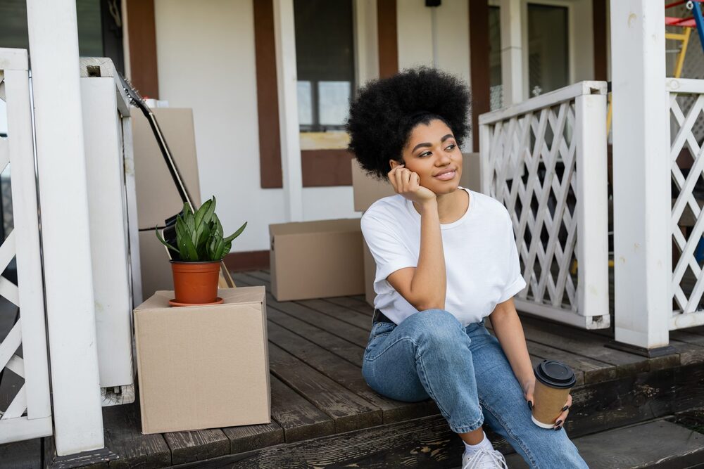A woman with curly hair sits on a porch with moving boxes and plants around her