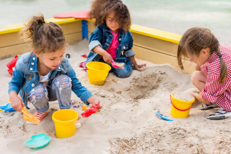 Three children play in a colorful sandbox, with one girl kneeling and holding a red toy, while another uses a pink shovel in a yellow bucket.