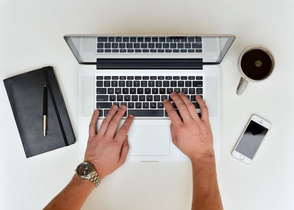 person using computer, with a notepad, cellphone and cup of coffee on the side