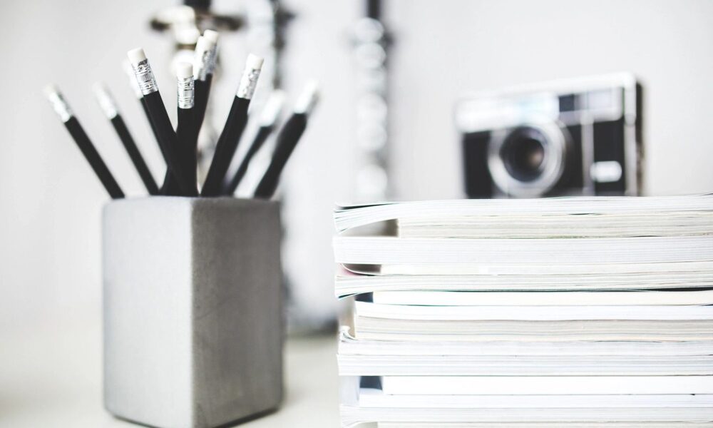 A concrete pencil holder with black pencils next to a stack of books and a blurred vintage camera.