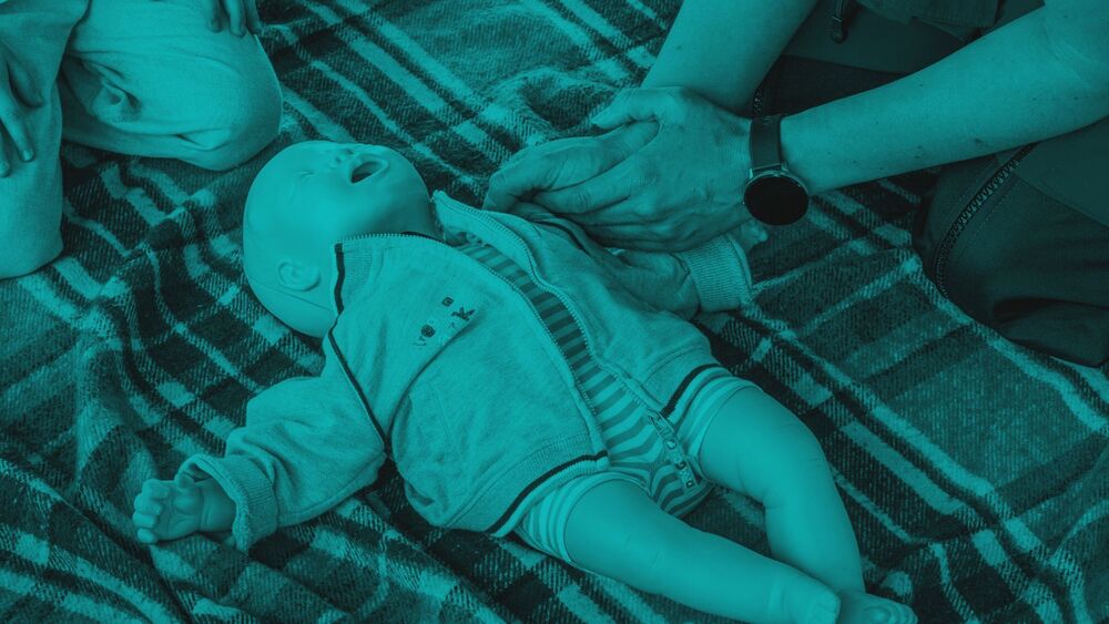 Hands performing CPR training on an infant mannequin lying on a plaid blanket, styled with a monochromatic teal overlay.
