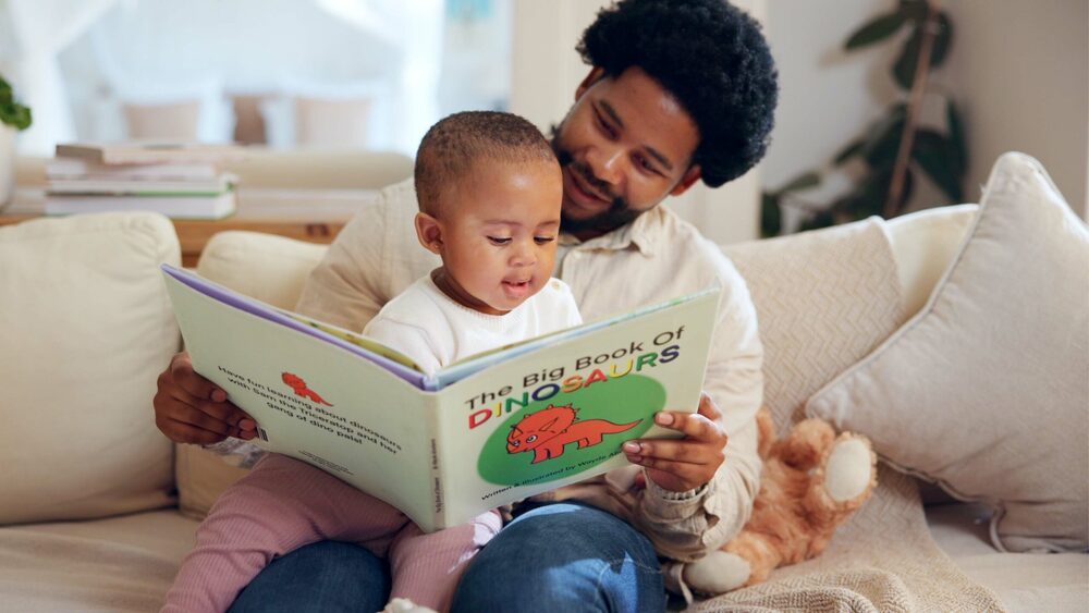Adult and young child sitting on a couch reading a large picture book together at home.