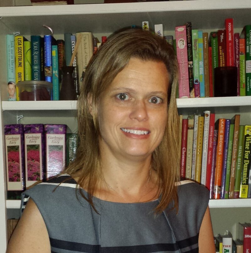 A woman stands in front of a bookcase filled with various books, smiling and looking towards the camera.