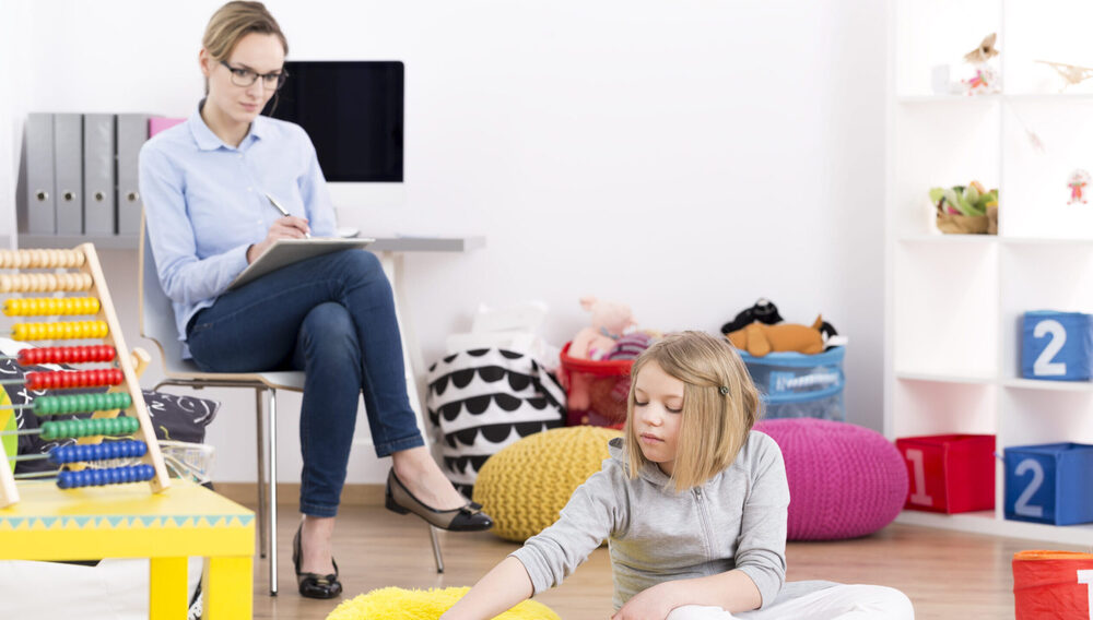 A child therapist taking notes on a clipboard while observing a young girl playing on the floor in a bright playroom."