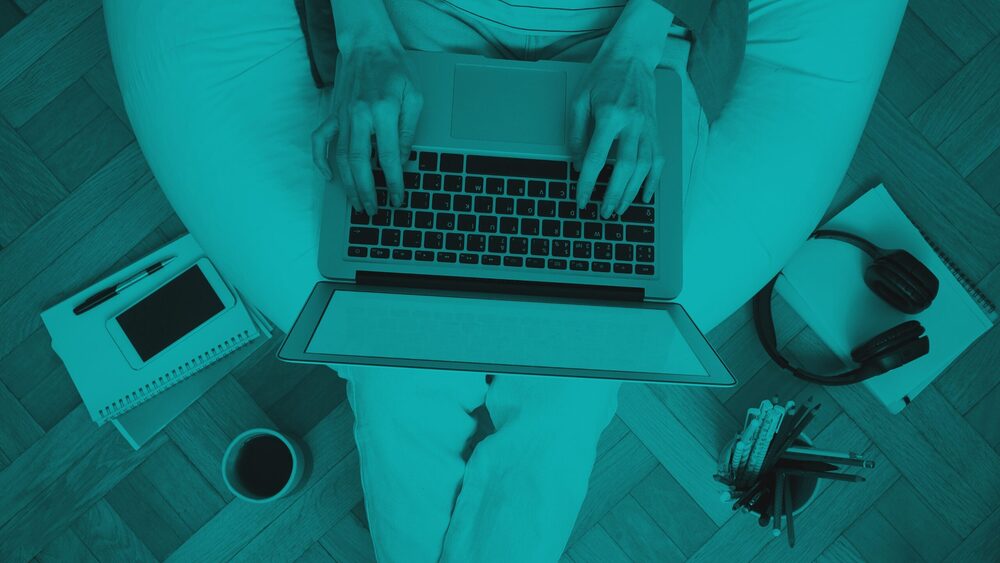 Top-down view of a person typing on a laptop while sitting on a beanbag chair, surrounded by a notebook, smartphone, headphones, and a cup of coffee, all overlaid with a teal tint.