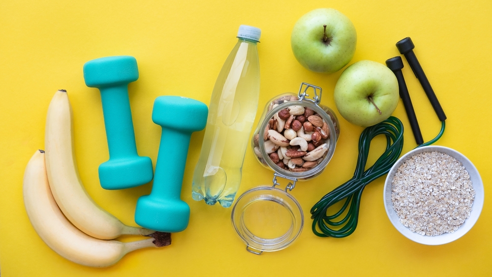 Assorted fitness and healthy lifestyle items on a yellow background, including bananas, green apples, dumbbells, a water bottle, nuts, a jump rope, and a bowl of oats.