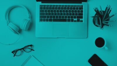 Top-down view of a workspace with a laptop, headphones, glasses, notebook, smartphone, coffee cup, and pencil holder, styled with a monochromatic teal overlay.