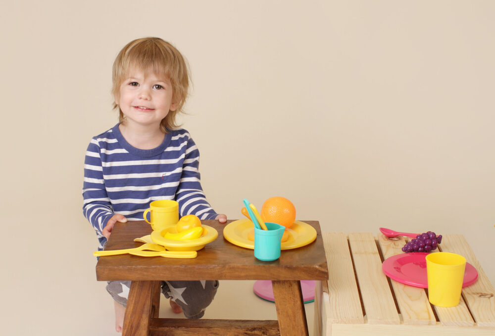 A smiling toddler playing pretend kitchen with colorful toy dishes and plastic fruit.