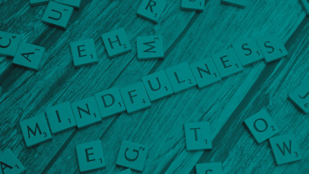 Scrabble letter tiles on a wooden surface spelling “MINDFULNESS,” with other scattered tiles nearby.