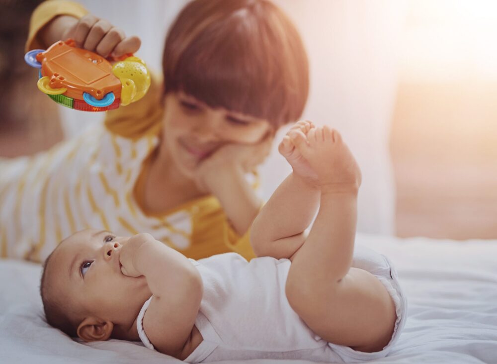 baby lays on its back with fist in mouth while older child holds a toy above its head