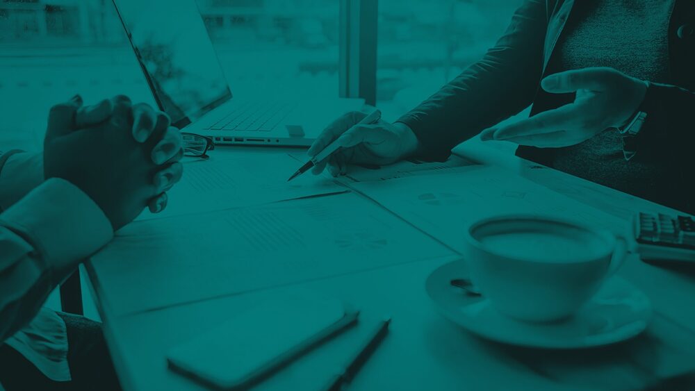 Two people review business documents and charts at a table with a laptop and a coffee cup, styled with a monochromatic teal overlay.
