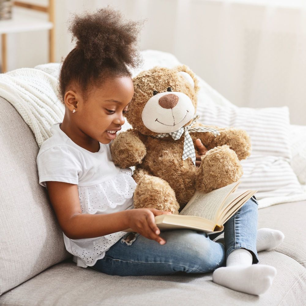prek girl sitting on sofa reading book with stuffed bear