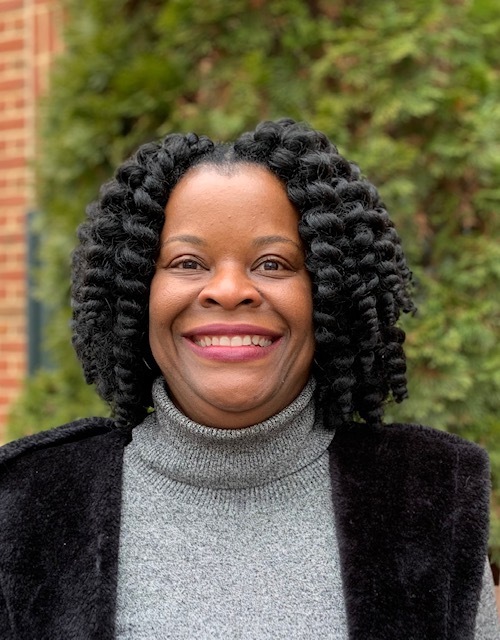 A woman with curly hair is smiling, wearing a grey turtleneck sweater and a black vest, standing outdoors in front of greenery and a brick wall.