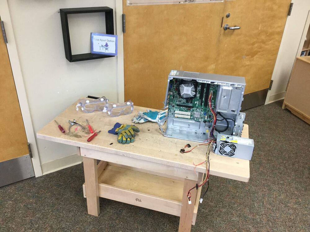 A classroom take-apart station with an open computer tower, safety goggles, gloves, and hand tools laid out on a wooden table.