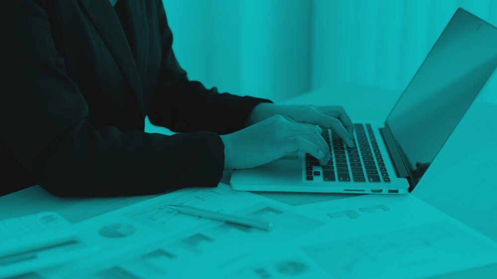 A close-up of a person in a blazer typing on a laptop at a desk with charts and graphs, styled with a monochromatic teal overlay.