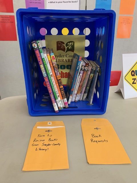 A blue plastic crate holding children's library books, sitting on a table alongside two manila envelopes labeled for book requests and forms.