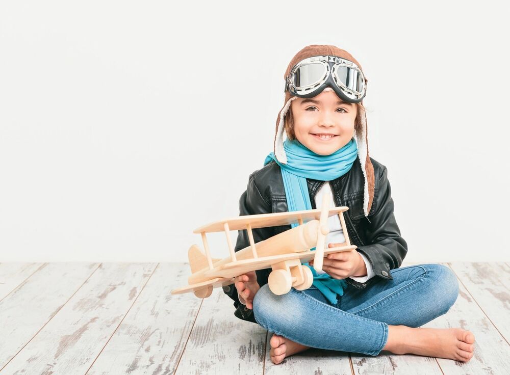 A child dressed as a pilot holding a wooden toy airplane on a white background.