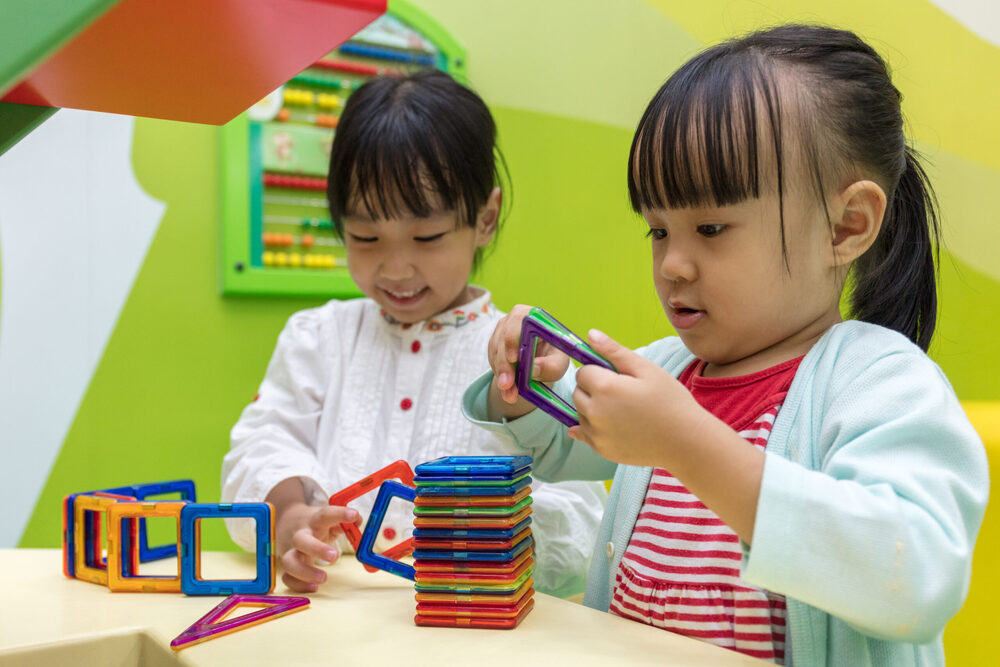 Two young black haired children playing with colorful toys in a green classroom