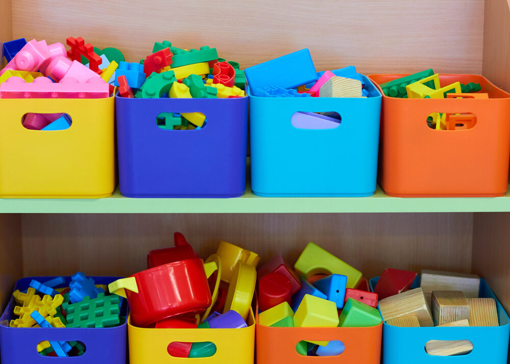 Color-coded storage bins filled with building blocks and toy cookware organized on a wooden shelf.