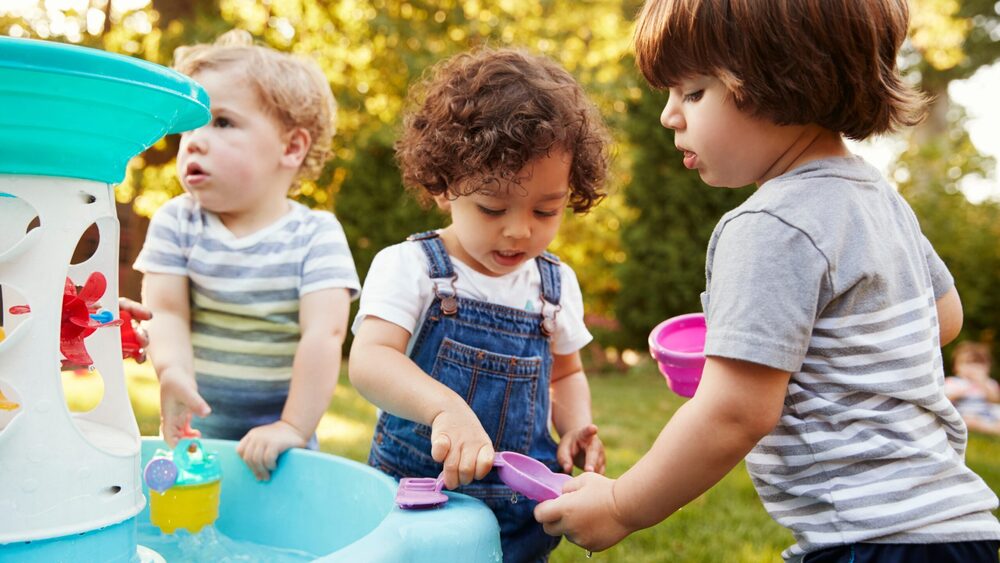 Three toddlers playing with scoops and cups at a water table outside in the sun.