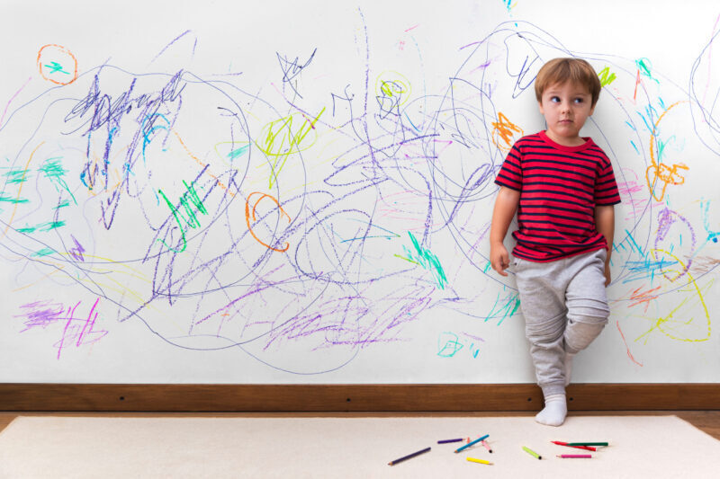 Child in red striped shirt stands in front of a wall full of colorful crayon scribbles