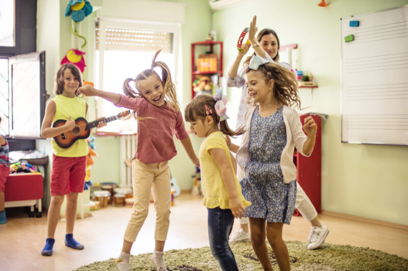 four children in a classroom dance while their teacher plays the tambourine